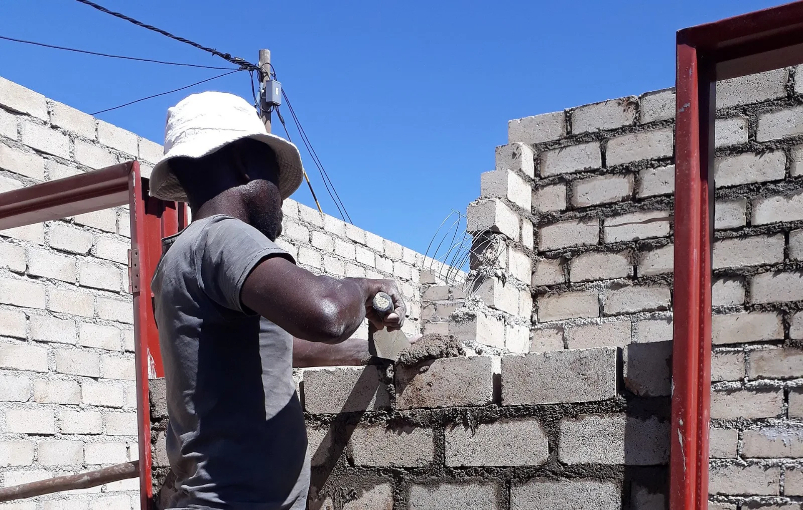 Zidar care întinde mortar cu mistria pe un perete din blocuri de beton, între tocurile metalice ale ușilor unei case în construcție.