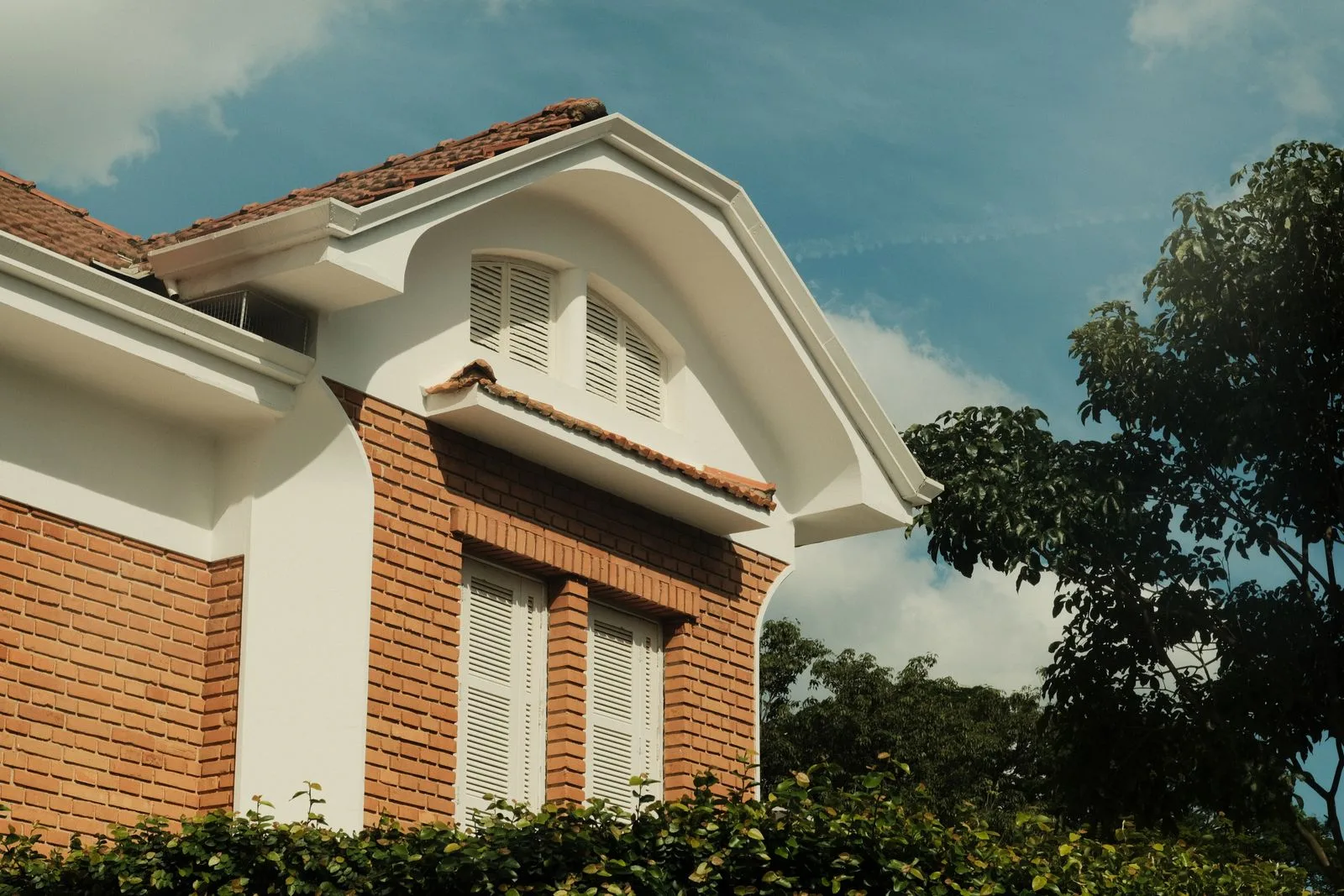 Fronton al unei case cu pereți din cărămidă aparentă, obloane albe și acoperiș din țiglă ceramică.