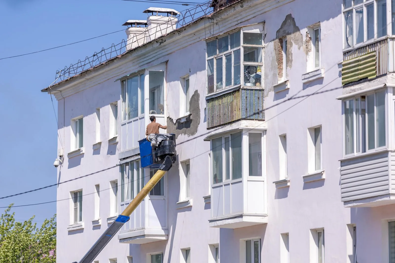 Muncitor într-o nacelă aplică tencuială pe fațada unui bloc alb cu mai multe etaje.