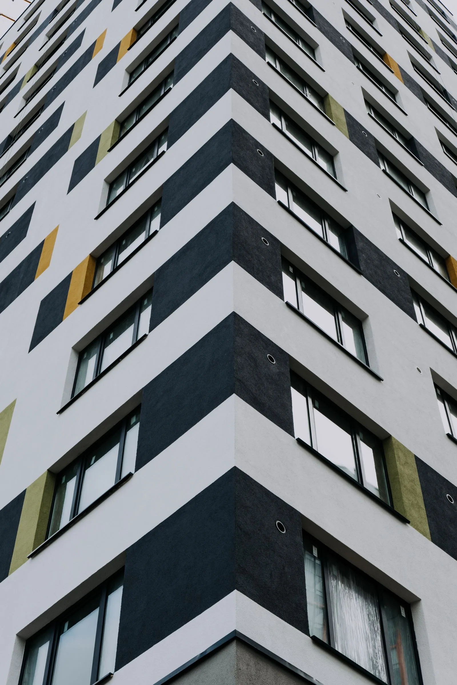 Colț de bloc înalt cu fațadă tencuită în benzi albe, gri închis și verde măsliniu, văzut de jos.