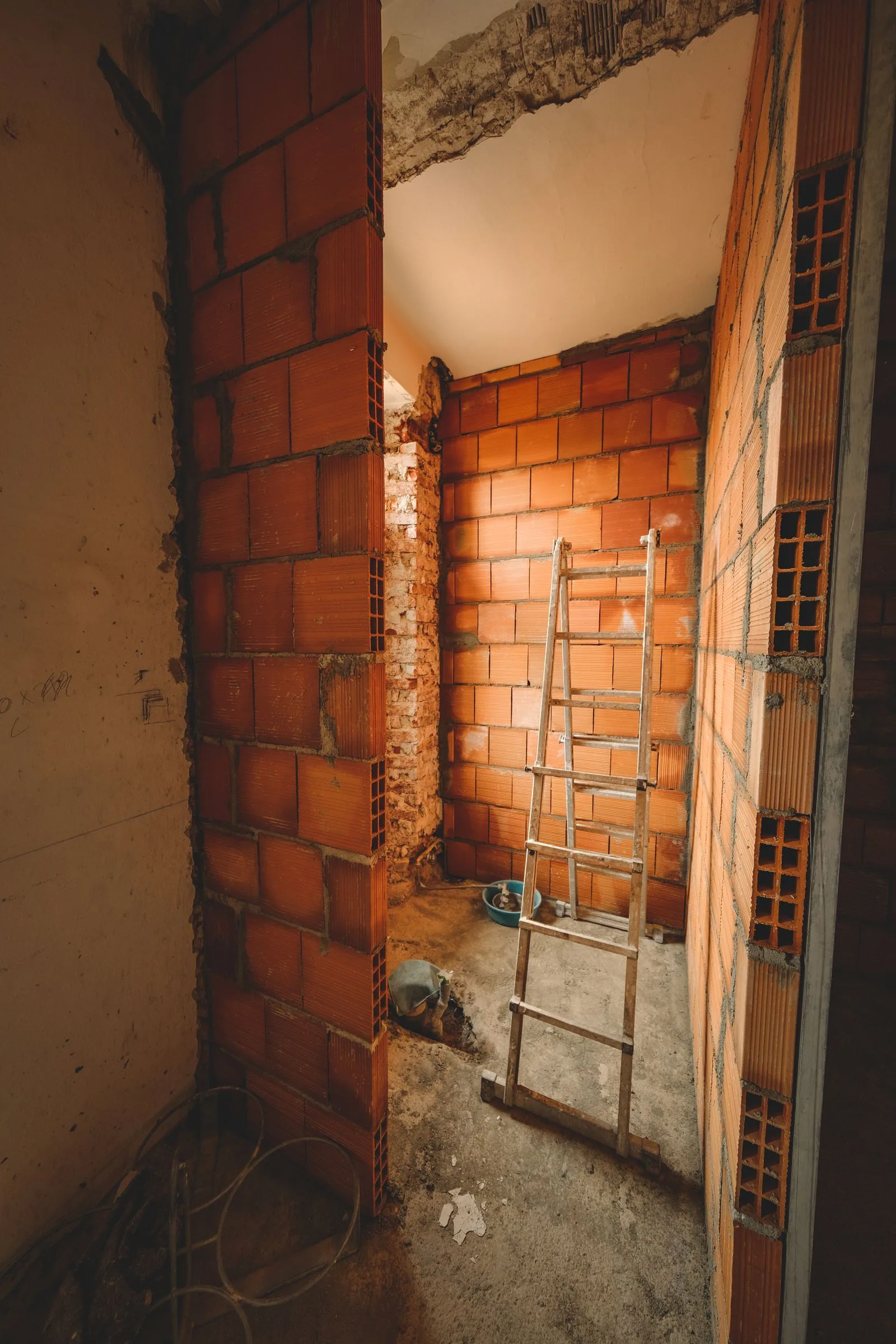 Cameră mică în reconstrucție, cu pereți noi din cărămidă cu goluri, o scară de lemn și pardoseală de beton.