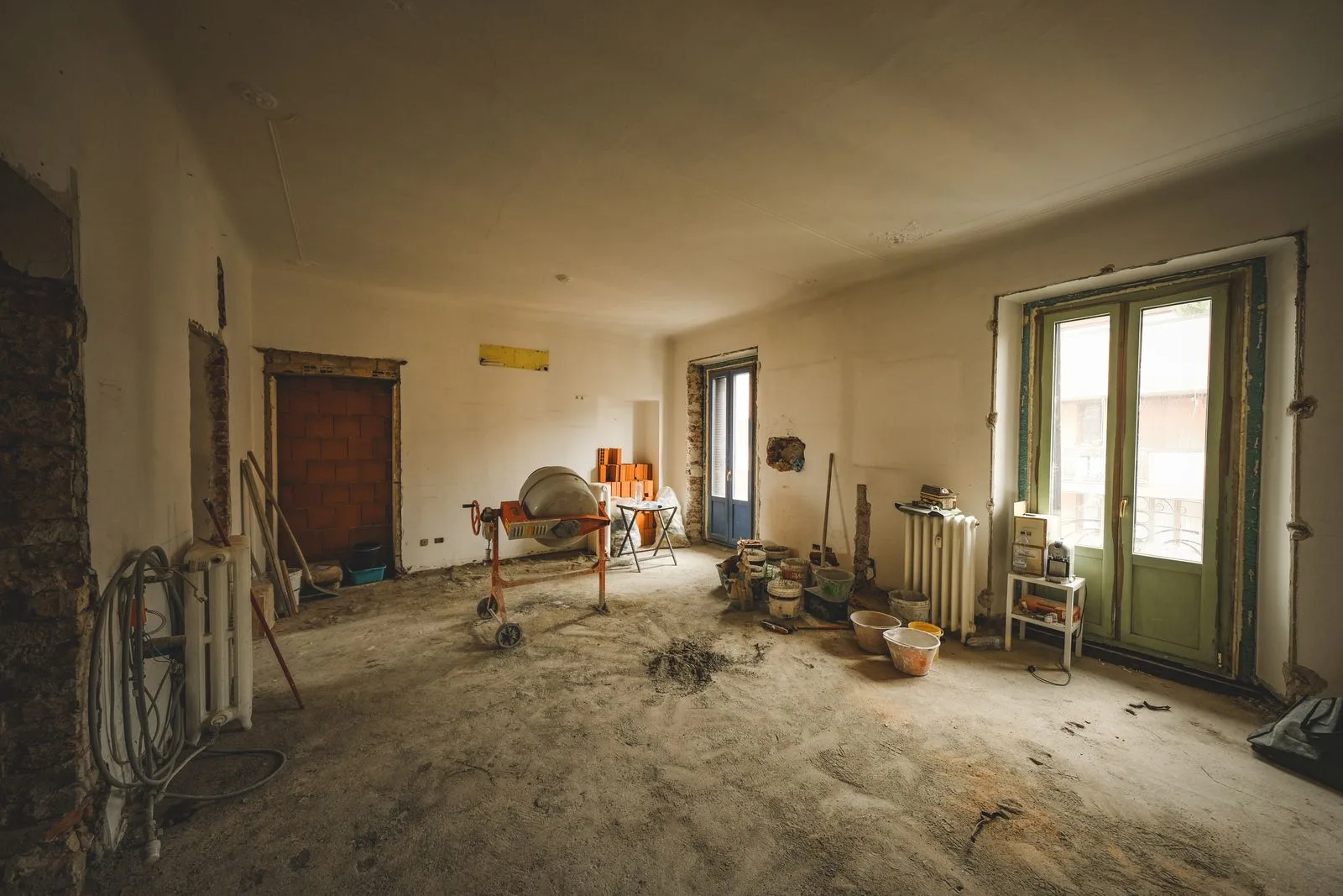 Cameră mare în renovare, cu betonieră, găleți, un perete decopertat și calorifere vechi.