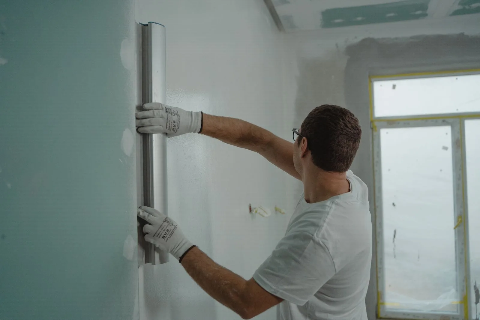 Muncitor văzut din spate verifică cu un dreptar un perete tencuit, într-o cameră în renovare.
