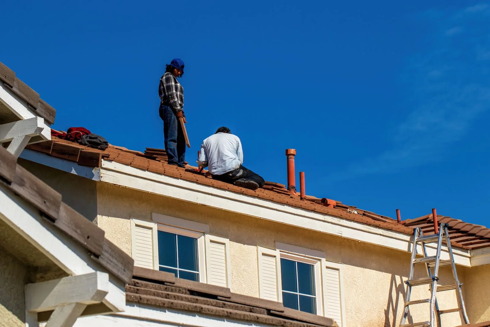 Doi muncitori repară un acoperiș de țiglă pe o casă, cu o scară sprijinită de perete.