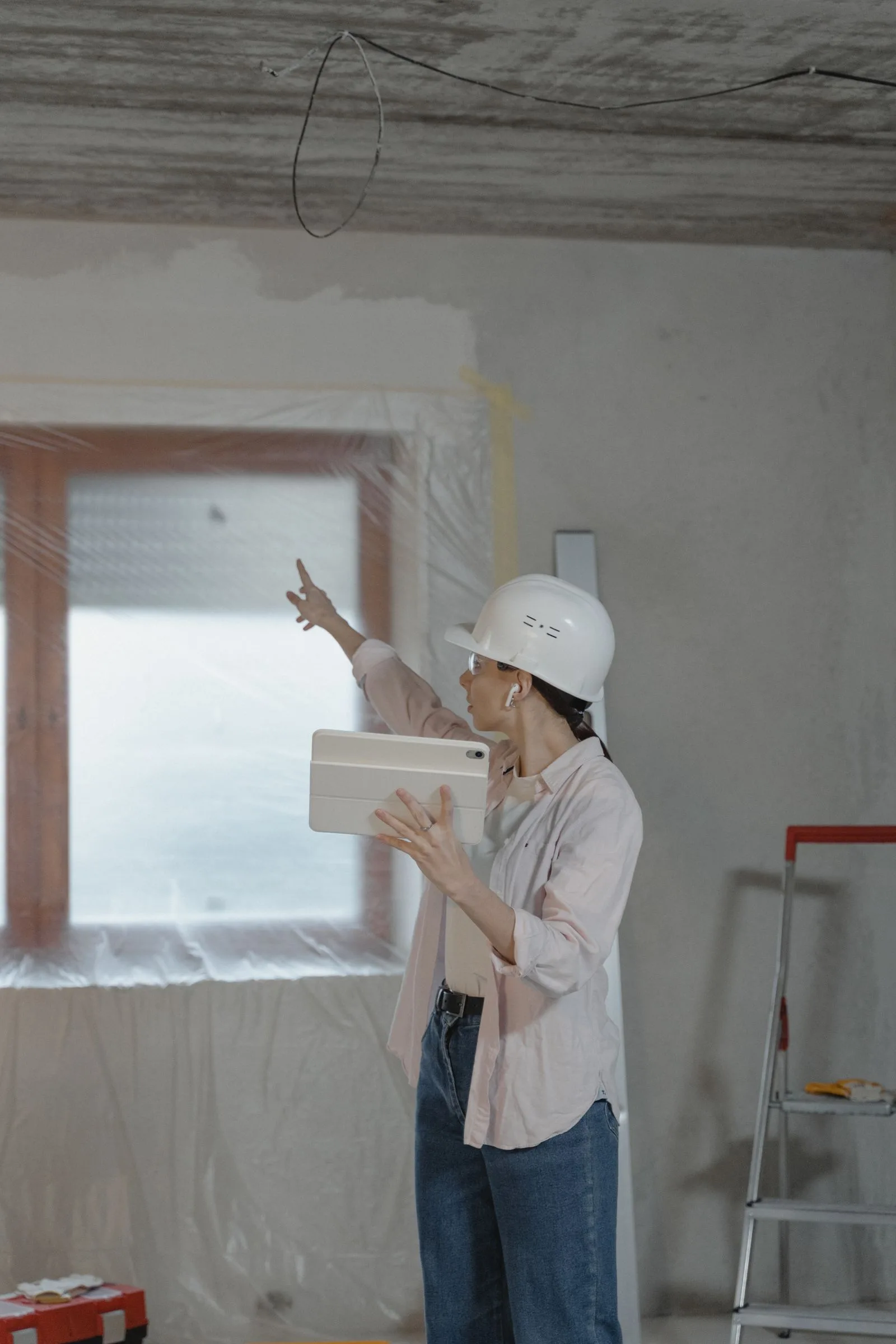 Persoană cu cască și tabletă indică o fereastră acoperită cu folie într-o cameră în renovare.