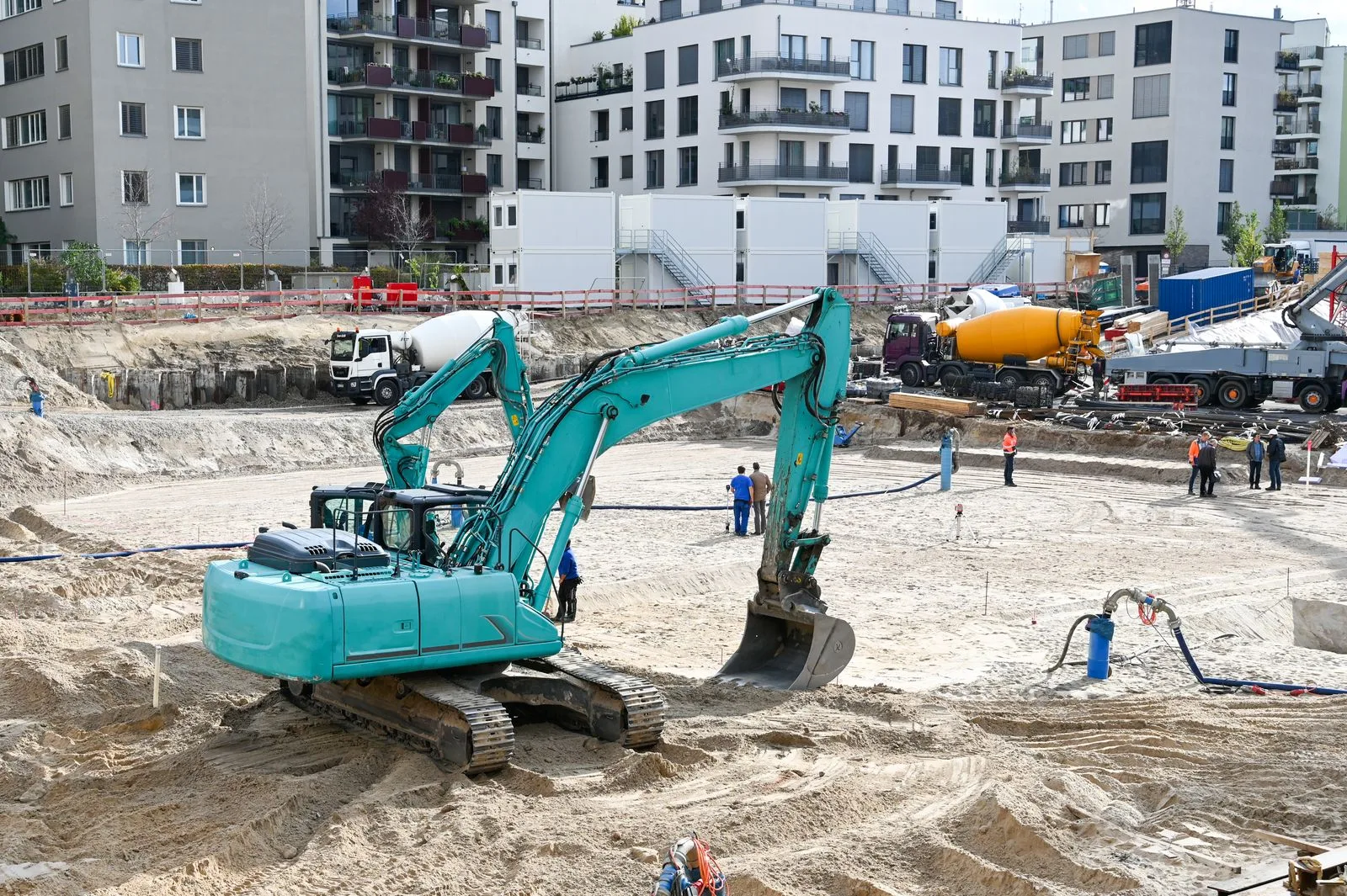 Excavator pe șenile lucrând într-o groapă de fundație largă, cu autobetoniere și blocuri în fundal.